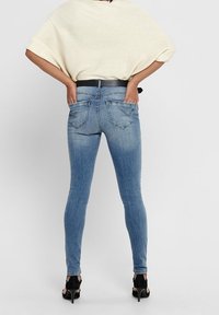 Femme portant un jean skinny bleu clair, un haut crème à manches courtes, une ceinture noire et des chaussures à talons hauts noires, debout avec les mains dans les poches arrière.