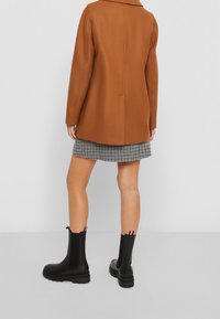 Bruine, oversized jas met een kraag, gedragen over een houndstooth rok en zwarte enkellaarsjes. De laarsjes hebben trekbanden met rode, witte en blauwe accenten.