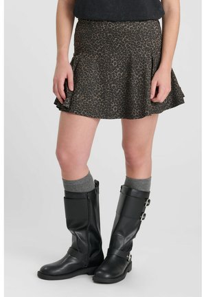 Person trägt einen kurzen braunen Rock mit Leopardenmuster, graue Socken und schwarze Stiefel bis zur Mitte der Wade mit Schnallen, steht auf einem einfarbigen Hintergrund.