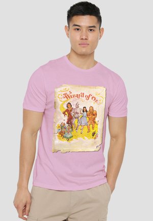 Man wearing a pink T-shirt with a vintage-style "The Wizard of Oz" design featuring Dorothy, Tin Man, Scarecrow, Lion, and other characters.
