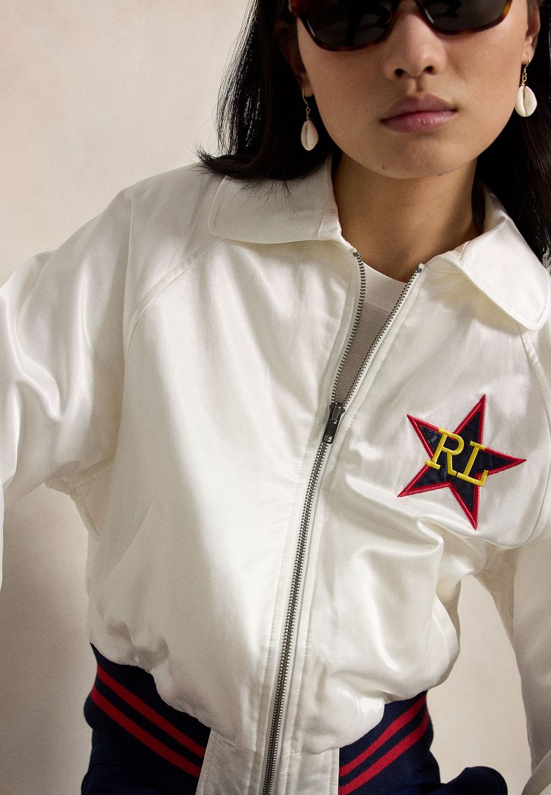 Femme portant une veste blanche zippée avec un écusson en forme d'étoile rouge et noire et les lettres RL, des lunettes de soleil foncées, et des boucles d'oreilles en coquillage sur un fond uni.