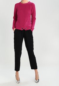 Frau trägt einen magentafarbenen Strickpullover mit Spitze, schwarze knöchellange Hosen und silberne High Heels, steht mit den Händen in den Taschen.