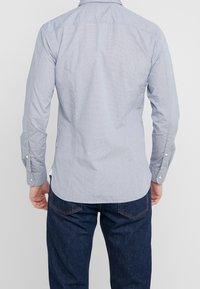 Hombre con una camisa de botones de manga larga azul clara y jeans azul oscuro, visto desde atrás contra un fondo liso.