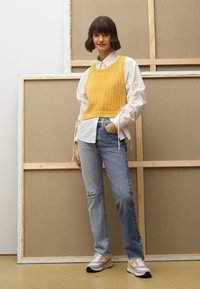 Geel gebreid sweater over een wit shirt, gecombineerd met lichtblauwe gescheurde jeans engrijze sneakers met gele accenten. Achtergrond is neutraal.