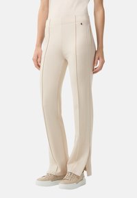 Pantalons beige ajustés avec une texture lisse, fente à la cheville et coutures verticales. Associés à des baskets beige clair pour le contraste.