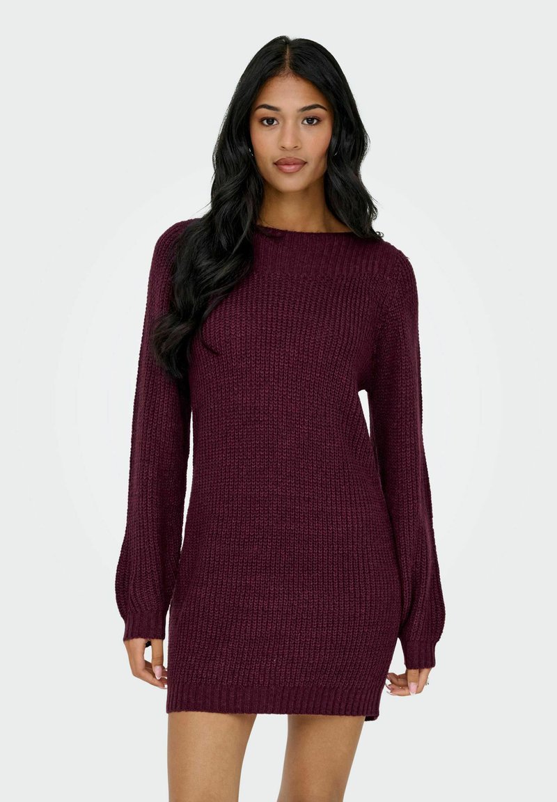 Donna con lunghi capelli neri che indossa un vestito maglione a maniche lunghe di color marrone bordeaux, in piedi davanti a uno sfondo chiaro e semplice.
