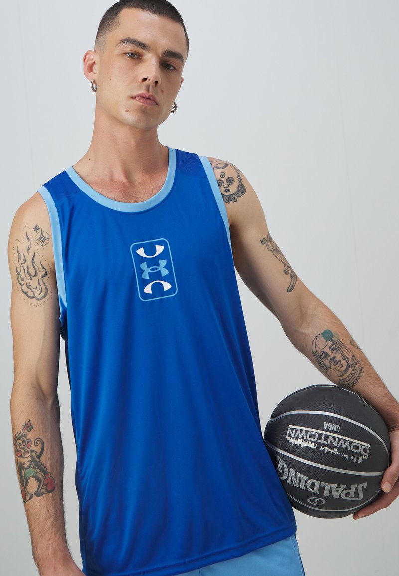 Niebieski sportowy top na ramiączkach z jasnoniebieskimi akcentami i logo z przodu, wykonany z gładkiego materiału. Model trzyma czarną piłkę do koszykówki.