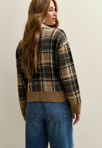 Cardigan en tricot avec un motif à carreaux marron, noir et crème, doté de poignets et d'un ourlet côtelés. Associé à un jean bleu ample.