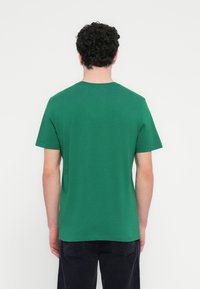 Πράσινο βαμβακερό μπλουζάκι με κοντά μανίκια και στρογγυλή λαιμόκοψη, εμφανιζόμενο από πίσω. Απλός σχεδιασμός, λεία υφή, χωρίς εμφανή λογότυπα ή μοτίβα.