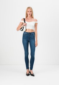 Top scurt alb, cu umerii la vedere și marginile ondulate, asortat cu blugi skinny negru închis, cu talie înaltă, și pantofi cu vârful ascuțit de culoare neagră.