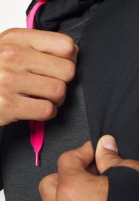 Ręce regulujące zamek w czarnej kurtce z widocznym jasnoróżowym sznureczkiem, skupione na akcji zapinania i detalach tkaniny.