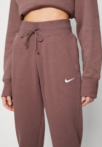 Bruna Nike joggers med dragsko i midjan, med sidofickor och en vit logotyp på låret. Mjuk tyg med ribbade ärmslut.