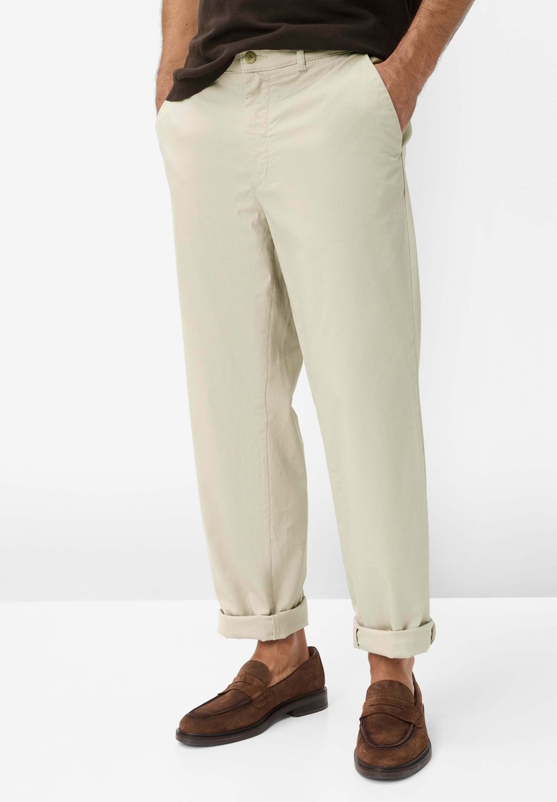 Mann trägt beige Chinos, an den Säumen hochgekrempelt, und braune Wildleder-Loafer, steht mit den Händen in den Taschen vor weißem Hintergrund.