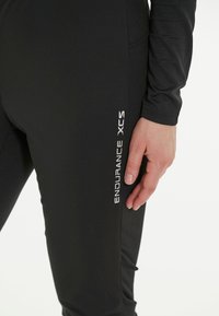 Mustat urheilulegginsit, joissa on sileä pinta, valkoisella "ENDURANCE XCS" -logolla sivussa, tiukka istuvuus ja kaareva sauma.