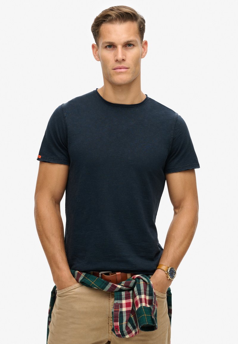 Camiseta de manga corta en color marino, fabricada en tela suave, con cuello redondo y una textura sutil. El modelo lleva un chaleco de franela a cuadros en verde y rojo atado a la cintura.