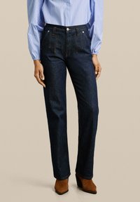 Donkerblauwe spijkerbroek met rechte pijpen, met voorzakken en contrasterende stiksels, gecombineerd met een lichtblauw gestreept blouse.