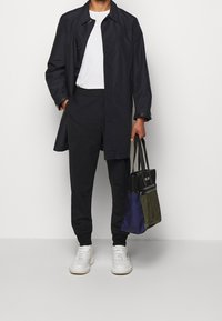 Granatowy, wodoodporny trencz na biały t-shirt, w zestawieniu z dopasowanymi ciemnymi spodniami i białymi sneakersami. Trzyma wielokolorową torbę na ramię.