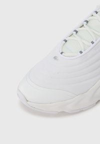 Λευκό αθλητικό παπούτσι με υφή από δίχτυ στο επάνω  μέρος, με ανοιχτό γκρι λεπτομέρειες και στρογγυλεμένη μύτη. Κορδόνια ενσωματωμένα σε μια δομημένη σχεδίαση.