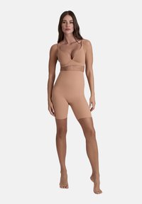 Donna in piedi a piedi nudi che indossa pantaloni modellanti a vita alta color carne e un reggiseno coordinato, su uno sfondo leggero e uniforme.