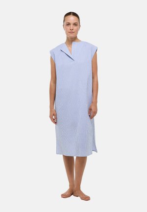 Femme debout pieds nus portant une robe bleu clair sans manches longueur genou avec un col asymétrique sur un fond uni.