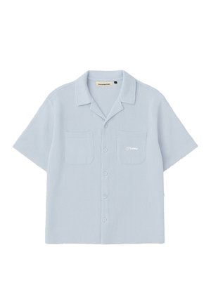 Femme portant une chemise bleu clair à manches courtes boutonnée et une casquette marine, regardant vers le bas, avec des boucles d’oreilles créoles dorées et les mains dans les poches.