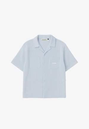 Frau trägt hellblaues kurzärmliges Hemd mit Knöpfen und eine marineblaue Kappe, schaut nach unten, mit goldenen Creolen und Händen in den Taschen.