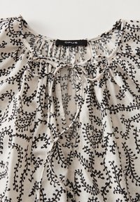 Lightweight blouse with a cream base, featuring black leaf patterns. Gathered neckline with a tie closure. Soft, flowing fabric.