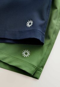 Dvi trumpų šortų poros mėlynos ir žalios spalvos audinyje, ant apatinio krašto yra saulės ženklas išsiuvinėtas balta siuvinėjimu. Lygus paviršius, pritaikyta konstrukcija.