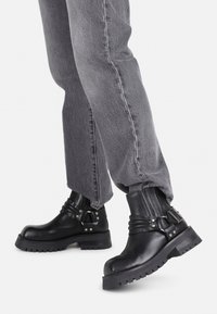 Botas de cuero negras hasta el tobillo con suela gruesa, detalles de hebilla y plisados verticales. Combinadas con jeans grises de corte tapered.