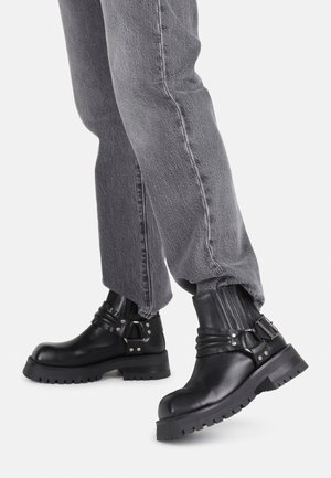 Botas de cuero negras hasta el tobillo con suela gruesa, detalles de hebilla y plisados verticales. Combinadas con jeans grises de corte tapered.