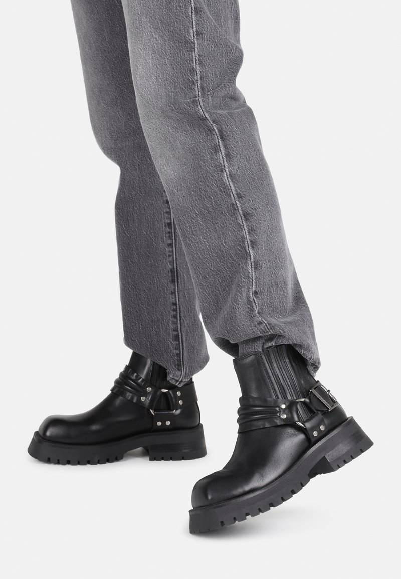 Botas de cuero negras hasta el tobillo con suela gruesa, detalles de hebilla y plisados verticales. Combinadas con jeans grises de corte tapered.