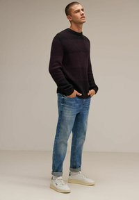 Tmavě pruhovaný pulovr s ribbovanou texturou, v kombinaci se světle modrými džíny rovného střihu a bílými teniskami s zeleným akcentem.