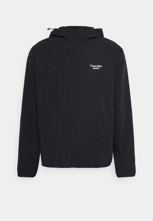 Veste coupe-vent noire à fermeture éclair avec capuche, manches longues, et logo blanc "Calvin Klein Jeans" sur le côté gauche de la poitrine.