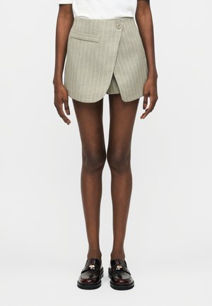 Μοντέλο που φοράει μπεζ μίνι φούστα με ρίγες και τύλιγμα μπροστά, λευκό τοπ και σκούρα καφέ δερμάτινα μοκασίνια, σε απλό φωτεινό φόντο.