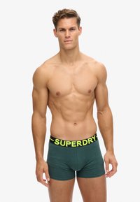 Calzoncillos boxer verde oscuro con una cinturilla negra que presenta el texto "SUPERDRY" en amarillo brillante. Tejido suave, diseño ajustado.