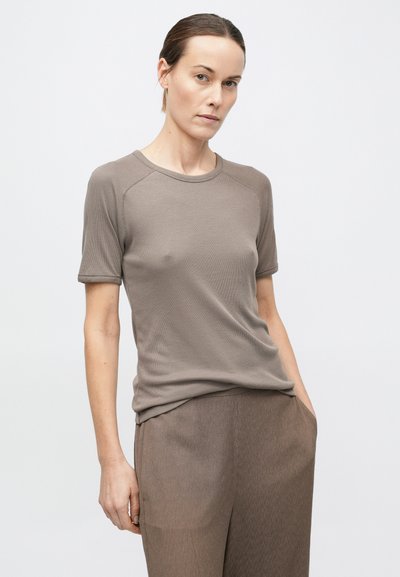 Sieviete ar gaišu ādu un aizpūstiem matiem, valkājot taupe krāsas īsām piedurknēm topu un atbilstošas vaļīgas bikses uz vienkārša fona.