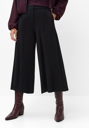 Femme portant un pantalon culotte noir à jambes larges, des bottes en cuir bordeaux et un haut bordeaux assorti avec manches, debout sur un fond blanc.
