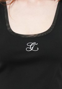 Juodas raukšlėtas marškinėlis su apvaliu iškirpimu ir permatomu puošimu, priekyje išsiuvinėta balta raidė "G".