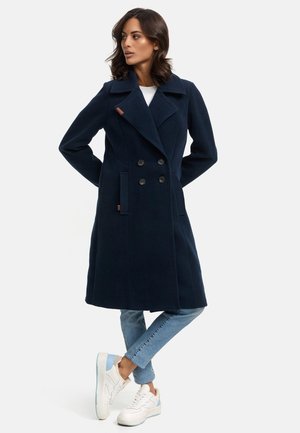 Navy wool-blend double-breasted coat with large lapels, two front pockets, and brown leather accent near shoulder. Worn over denim and sneakers.