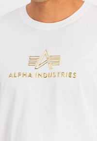 Bílý bavlněný tričko s pozlaceným reliéfním logo "ALPHA INDUSTRIES" a dekorativními liniemi v geometrickém vzoru na přední straně.