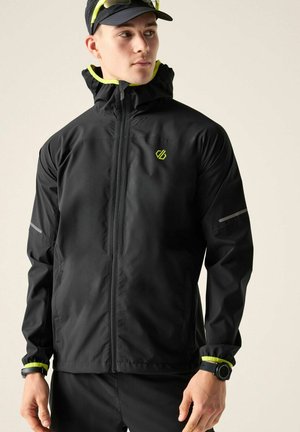 Junger Mann trägt eine schwarze Kapuzenjacke mit neongelben Akzenten, eine schwarze Kappe und eine schwarze Smartwatch und schaut seitlich vor einem einfarbigen Hintergrund.