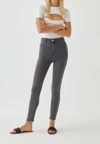 Jean skinny gris taille haute avec une texture lisse, associés à un haut blanc à côtes avec un design à découpe et des sandales tissées noires.