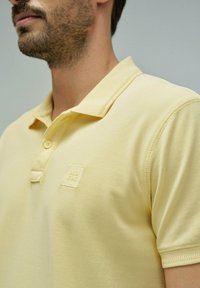 Hombre con una camiseta polo de color amarillo pálido con un pequeño logo de "SALSA JEANS" en el pecho, sobre un fondo liso.