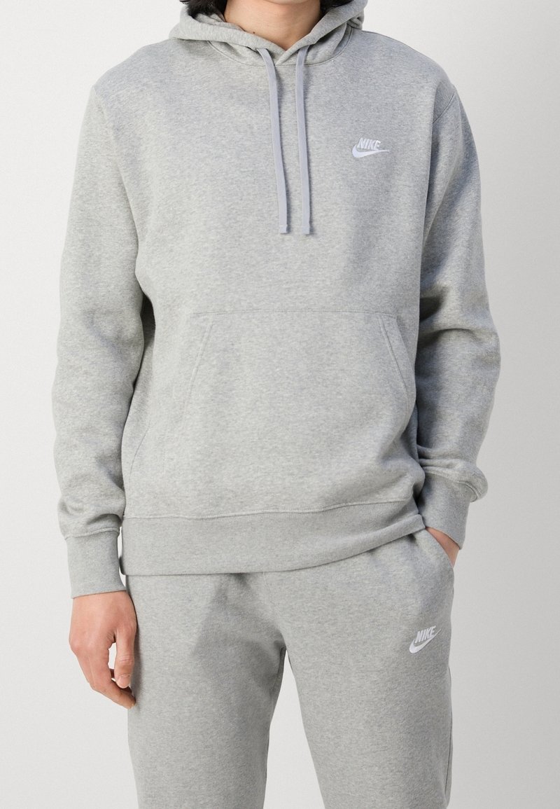 Person som bär en ljusgrå Nike-hoodie och matchande träningsoverallbyxor med vita Nike-logotyper, en hand i fickan, mot en enfärgad bakgrund.