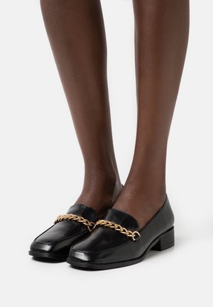 Mocassins en cuir noir avec un accent en chaîne dorée, talon bas, bout carré et texture lisse. Présentés avec des jambes dans une pose neutre.