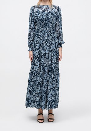 Femme portant une robe bleue à manches longues, longueur cheville, à motif paisley avec taille smockée et sandales noires à bout ouvert, debout devant un fond uni.