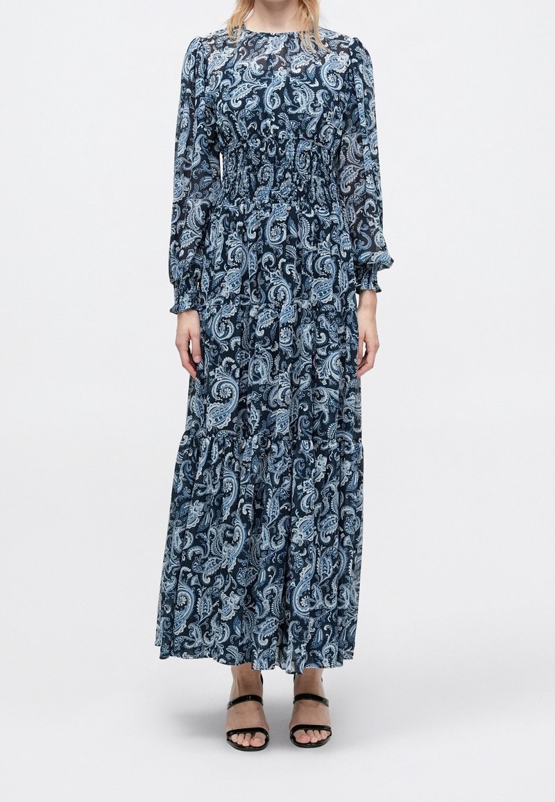 Femme portant une robe bleue à manches longues, longueur cheville, à motif paisley avec taille smockée et sandales noires à bout ouvert, debout devant un fond uni.