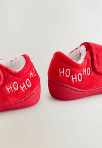 Punaiset tossut, joissa on pehmeä ja pörröinen kangas, valkoinen karvainen vuori ja brodeerattu "HO HO HO" -teksti sivuilla. Pyöreä muotoilu ja tasapohja.