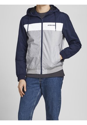 Mand iført en navyblå, lysegrå og hvid Jack & Jones hættetrøje med lynlås og hætte, kombineret med blå jeans.