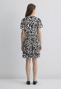 Kurzes, �ärmelloses Kleid mit einem schwarz-cremefarbenen Blattmuster, das eine geraffte Taille und einen geschichteten, gerüschten Saum aufweist. Glattes Material, lässige Passform.